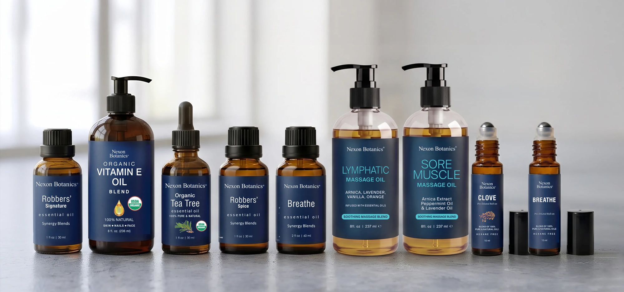 NexonBotanics®  Home of pure natural plant based products lineup of Robbers Signature essential oil and massage oil products on a light gray surface with a blurred white background. The products include amber glass bottles, clear bottles with amber oil, and black pump/dropper tops, with labels such as Robbers Signature, Vital E Oil, Tea Tree, Breathe, Lymphatic Support, Sore Muscle Massage Oil, Clove, and Breathable.