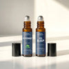 Two amber glass roll-on bottles with navy labels from Nexon Botanics, one labeled Peppermint and one labeled Zen Sleep, standing upright on a white surface with their black caps placed beside them in a bright, sunlit setting.